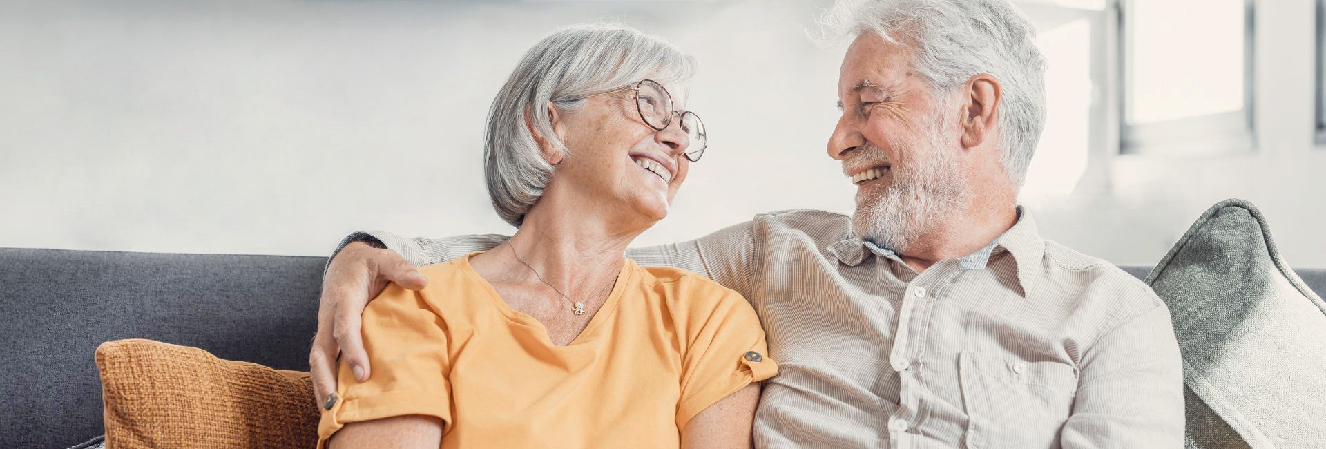 Älteres Paar sitzt eng beieinander auf einem Sofa, schaut sich zugewandt an und wirkt entspannt im häuslichen Umfeld