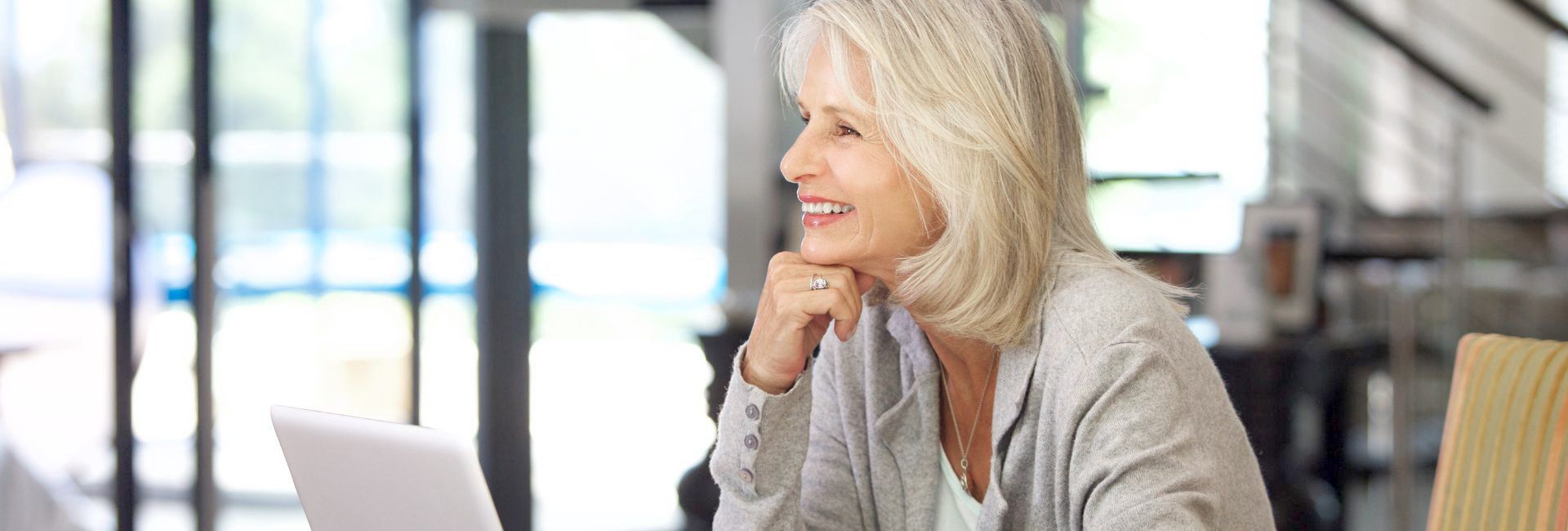 Eine ältere Frau sitzt an einem Tisch und blickt lächelnd aus dem Fenster, neben ihr steht ein Laptop